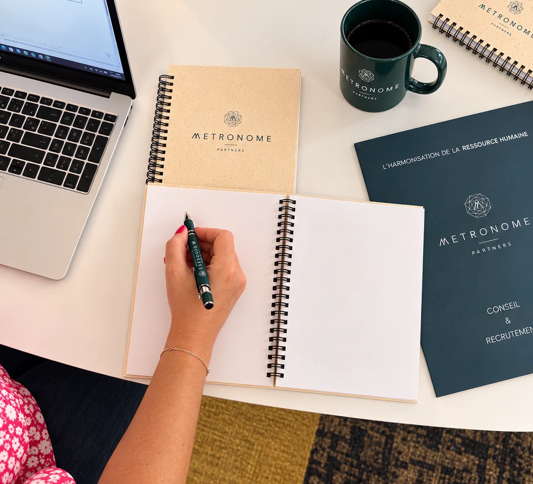 Une main écrit dans un carnet, entourée de carnets, d'un mug et d'une plaquette Metronome Partners